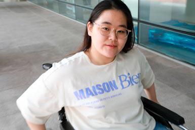 Flicka i tonåren sitter i en rullstol. Flickan med mörkt rakt hår, glasögon vit T-shirt med text i blått Maison Bien.